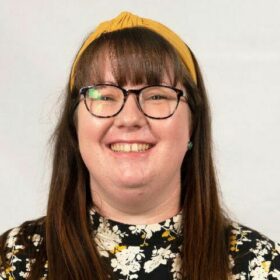 Sarah Wilkinson, a white lady with dark brown, straight hair and a fringe, wearing black glasses, a yellow headband and flowery shirt. She's smiling widely.