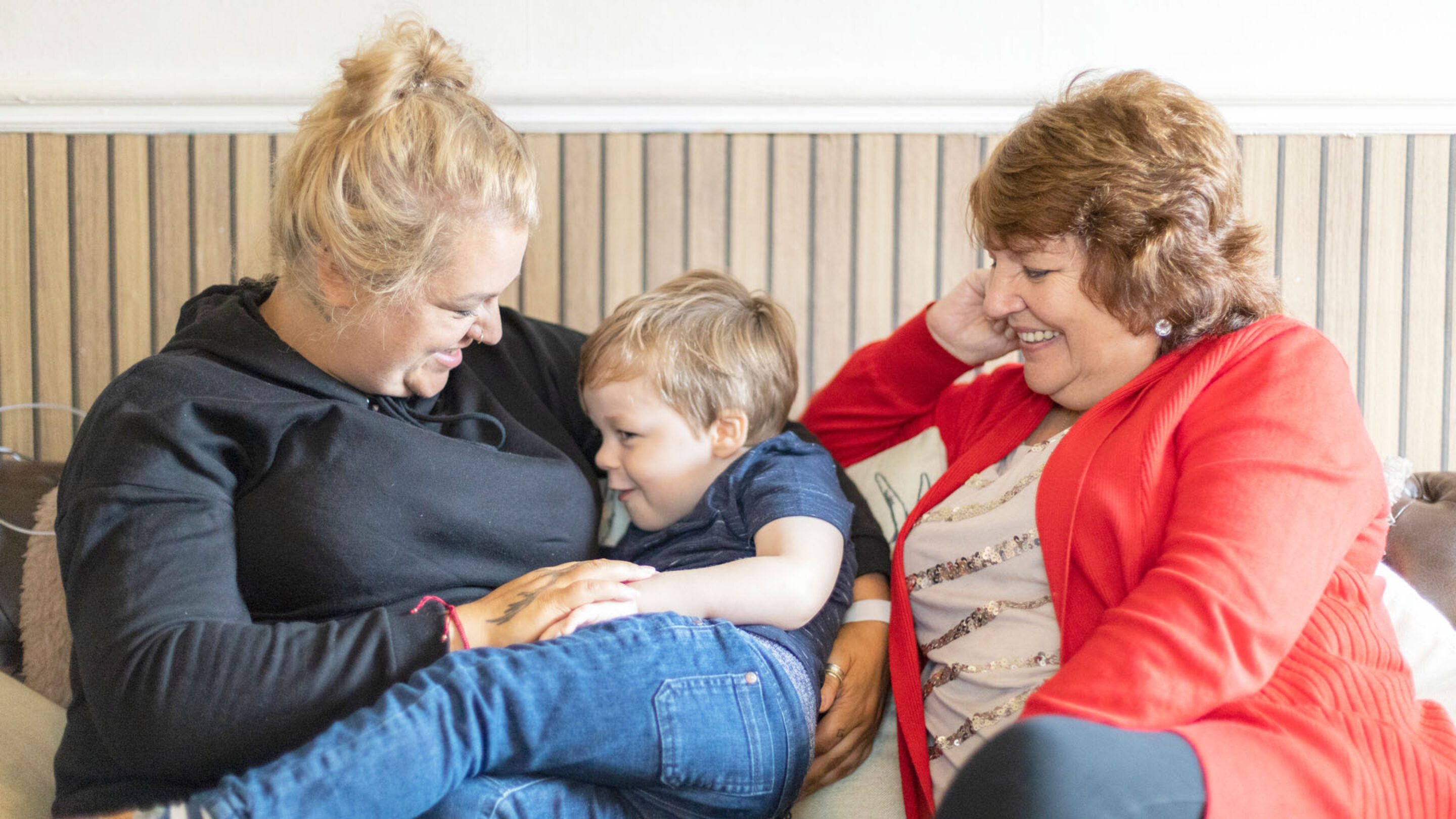 A mother sitting down and smiling whilst cuddling her son, her female friend is sat next to them