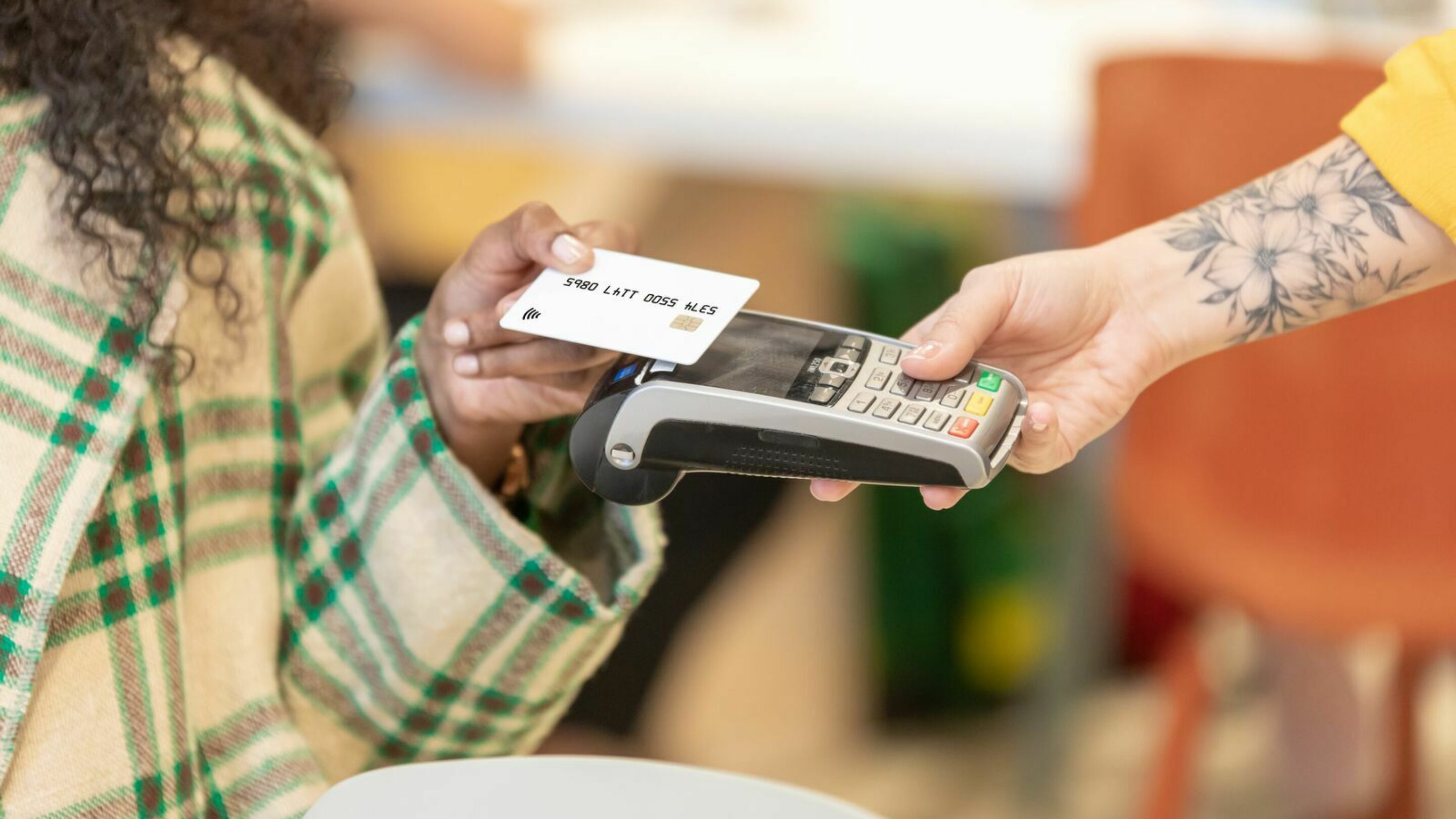 One lady, who is black with long black curly hair in a green chequered jacket, holds out her hand with a card in towards the card reader the white lady with tattoos and yellow jacket is holding towards her