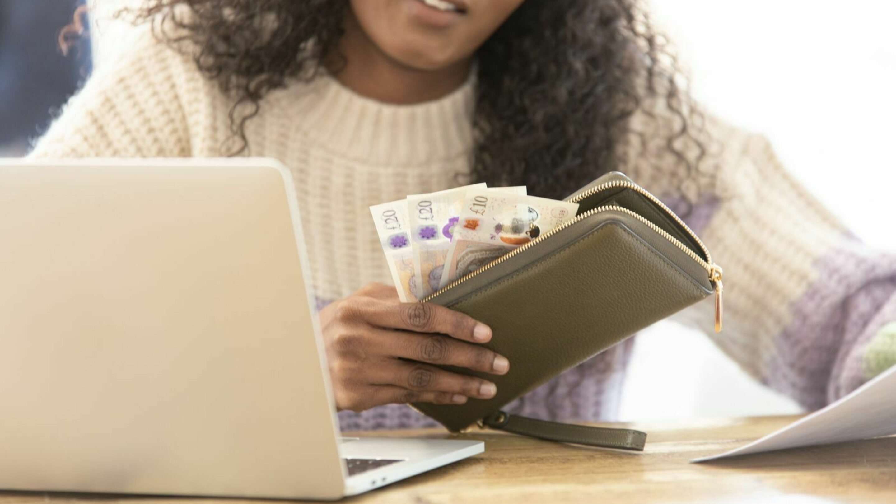 A young black lady with long curly hair, sat at a wooden dining table with her laptop and wallet containing cash, and holding a piece of paper.
