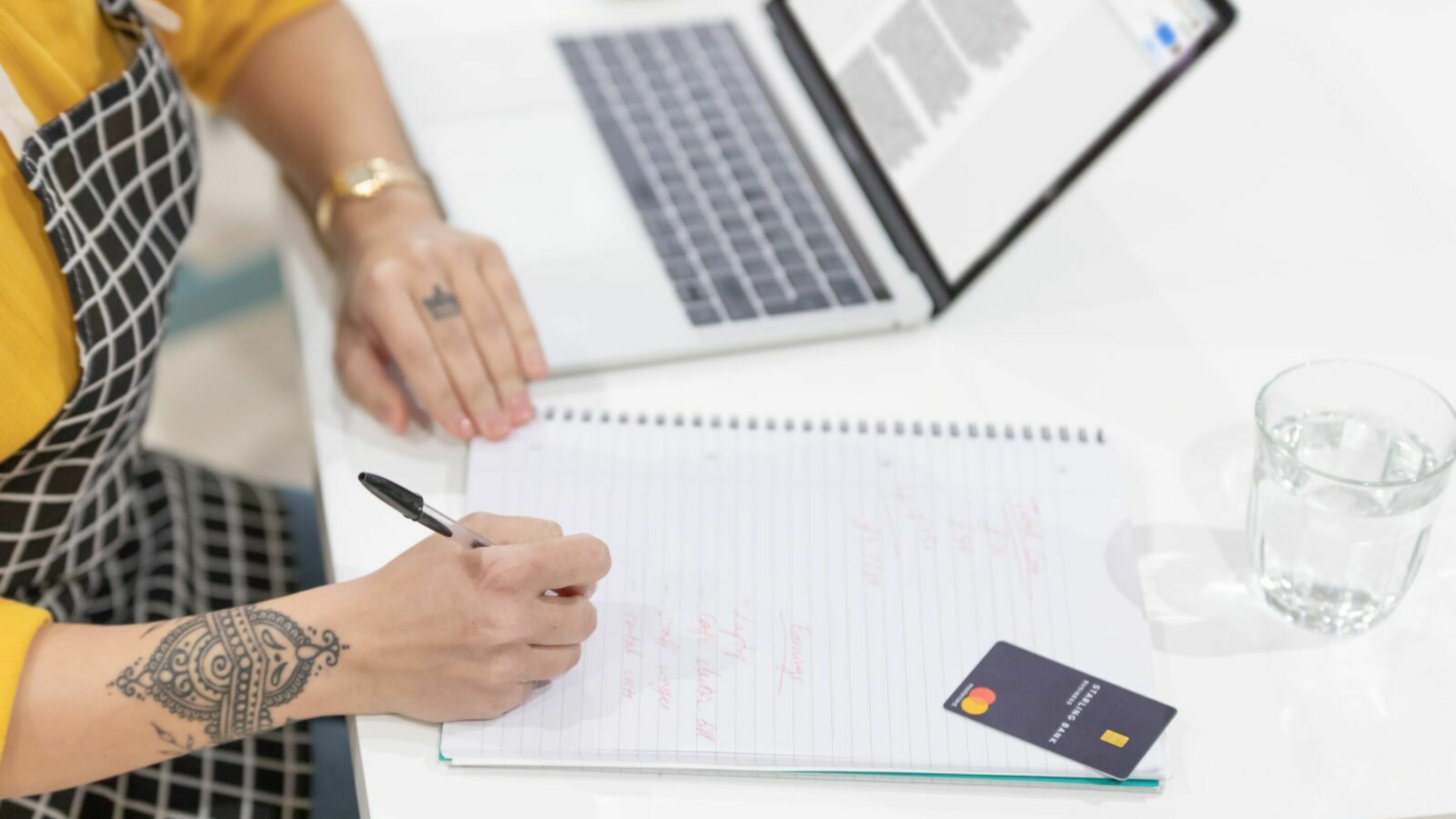 A woman with tattoos sat at a desk side on, with a laptop open, glass of water, a payment card and writing notes into a notebook