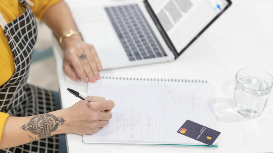 A woman with tattoos sat at a desk side on, with a laptop open, glass of water, a payment card and writing notes into a notebook