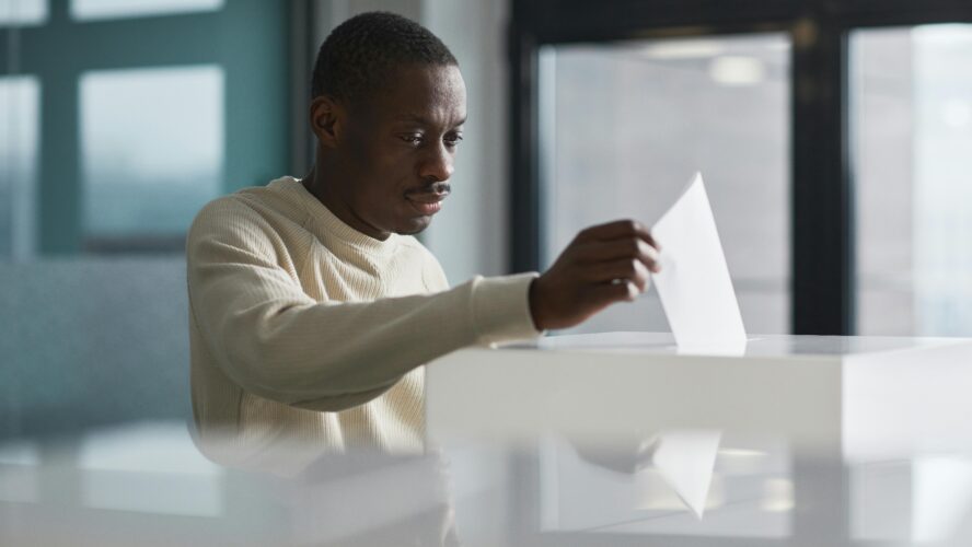 Black man in cream jumper at the polling station, placing his vote into the ballot box,