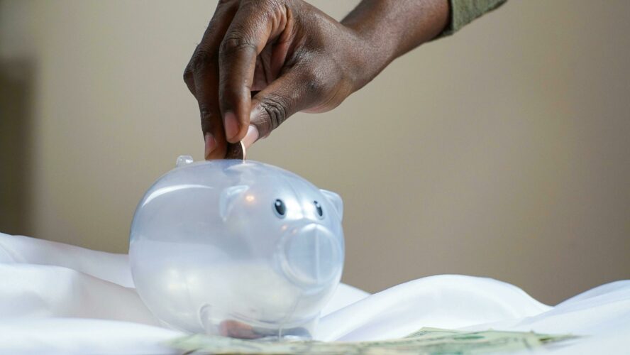 A black person's hand reaching to put a coin in a translucent white piggy bank. Photography owned by Joslyn Pickens.