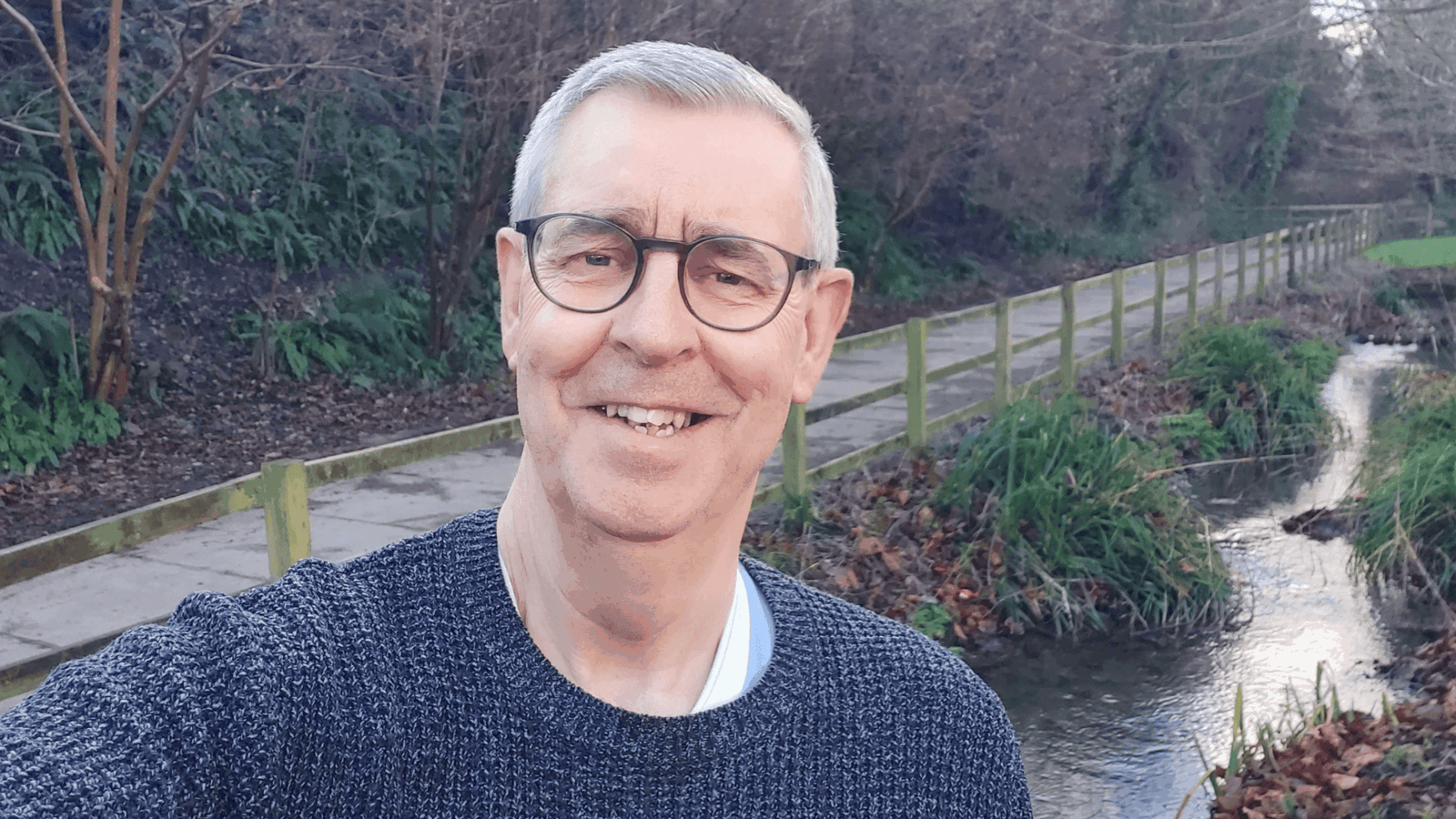Steve Hart, former Debt Coach of 10 years who is Chair of the CAP Management Committee in Yeovil. Steve is a white male with short grey hair, round black glasses and a navy jumper. Steve is stood next to a stream in a park or woodland area, smiling at the camera for a selfie.