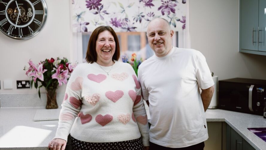 Angela, a white female with shoulder length brown hair, laughing next to her husband, a white male with no hair. He is also smiling.