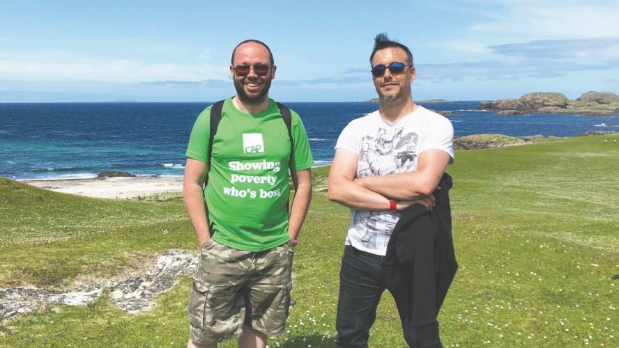 Two males stood with their arms folded smiling at the camera, in the background is the ocean.