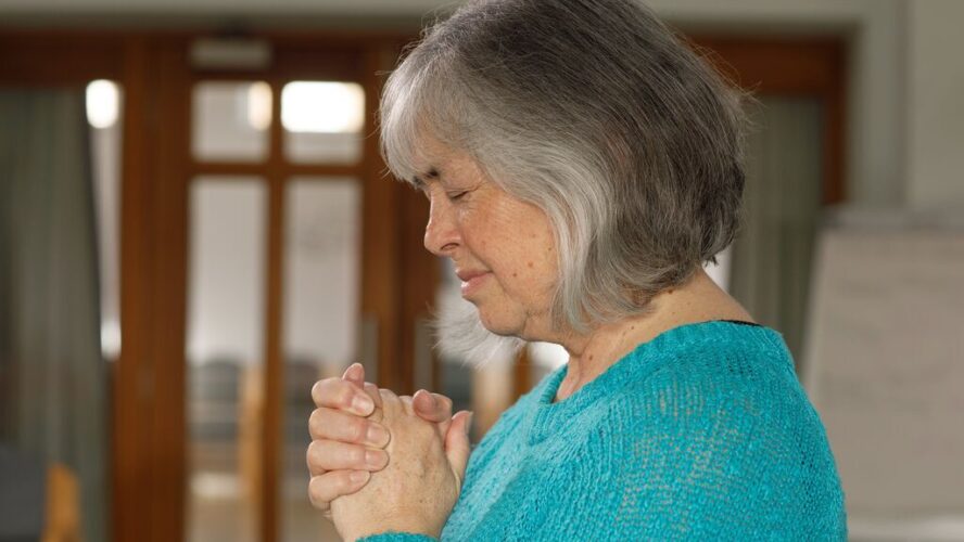 Side photo of a lady stood in a church with her head bowed slightly and her hands crossed, praying