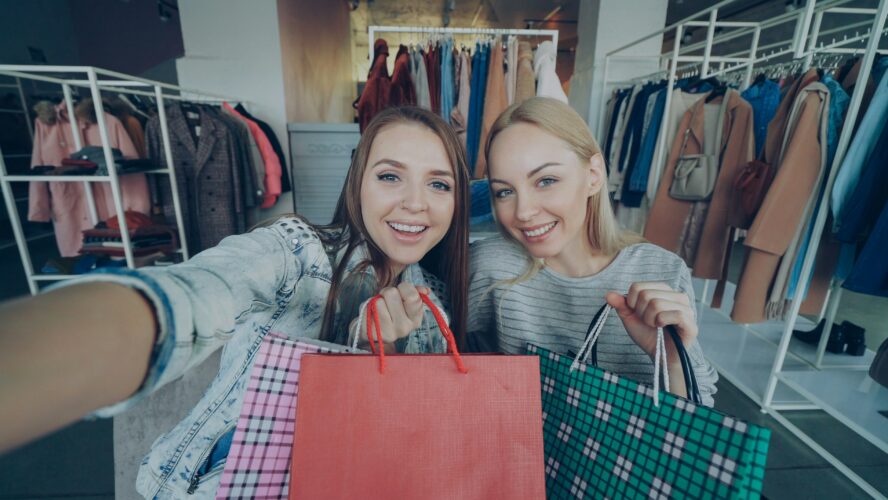 Two friends taking a selfie smiling, holding up shopping bags.