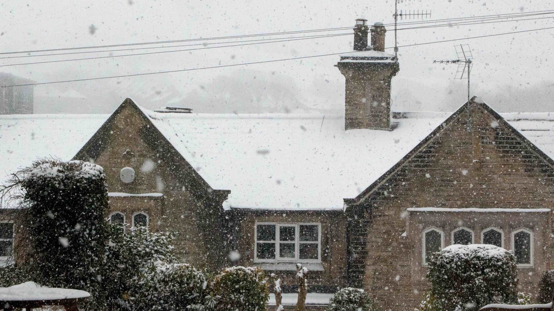 A wintery landscape of a large house with a chimney covered in snow, there are picnic tables in front.
