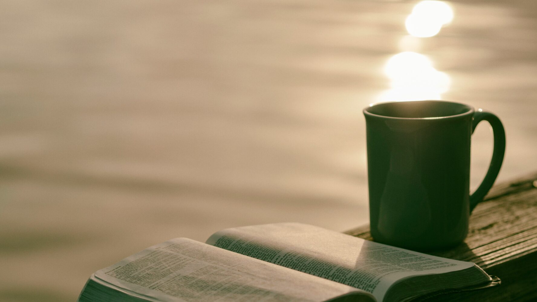 Image of an open Bible on a bench, next to a mug and a lake