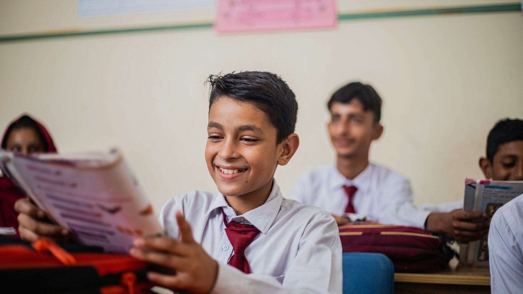 Image of a pupil sat in a classroom wearing a school uniform, smiling at their textbook