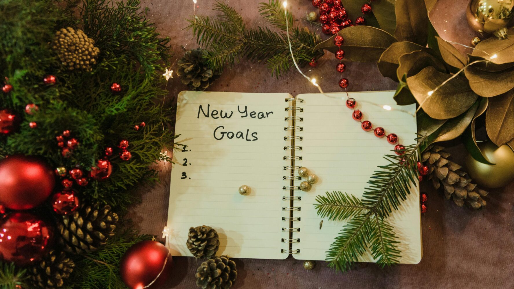 A notebook open with the words 'New Year Goals' at the top, on a table surrounded by Christmassy decor. Photo by Arina Krasnikova.