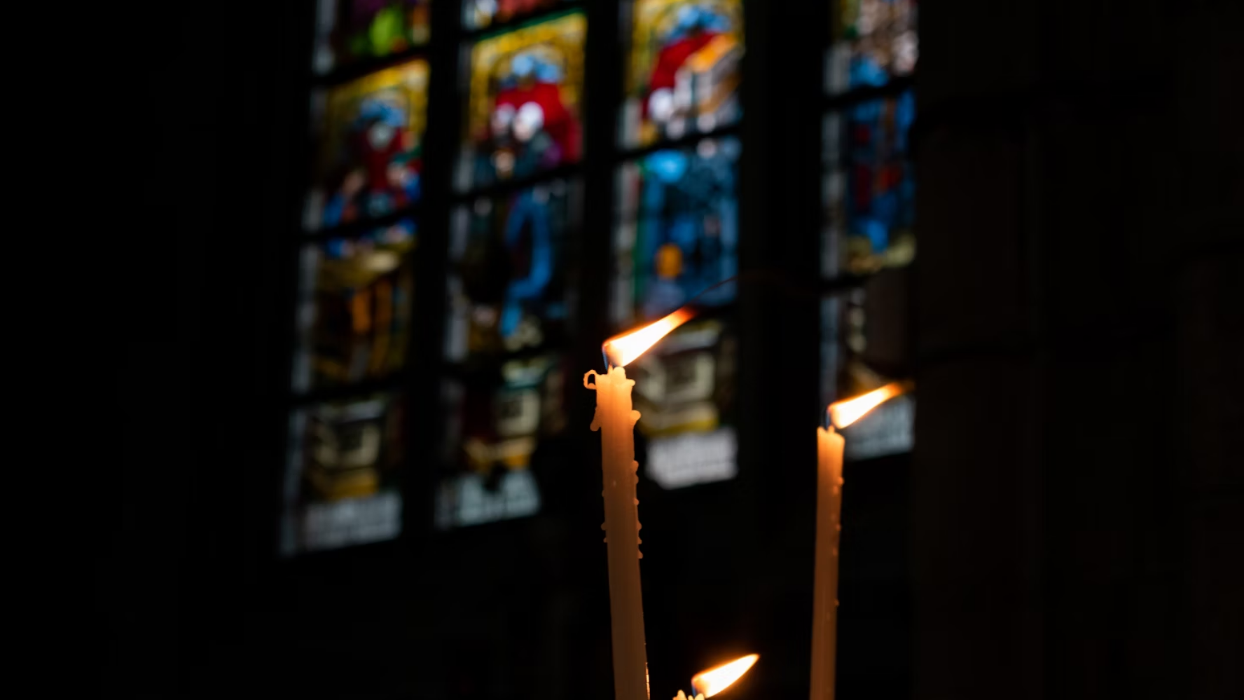 Three pillar candles lit in a dark traditional church, with colourful stained glass behind them