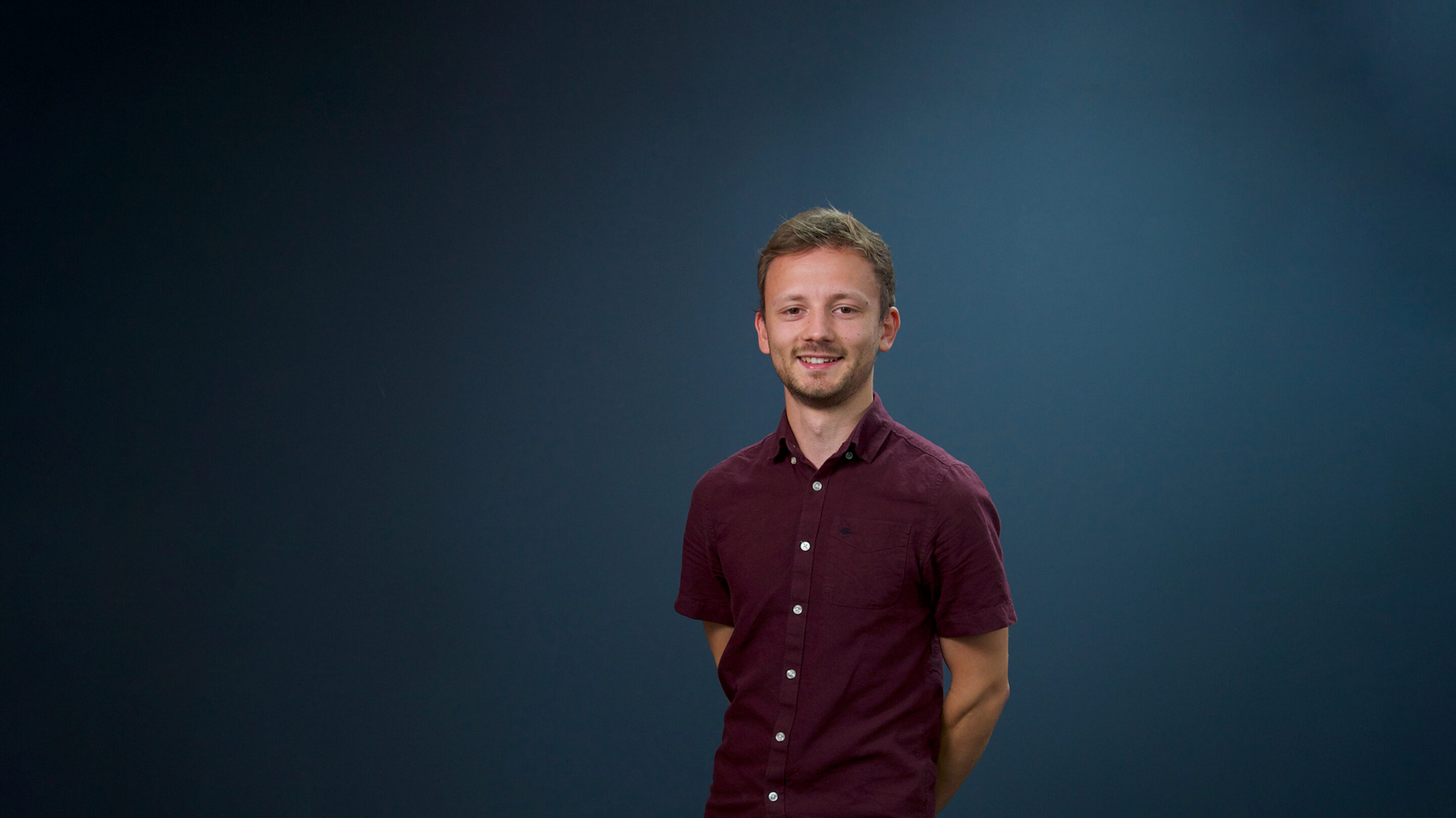 Young male stood with his hand behind his back looking towards the camera and smiling.