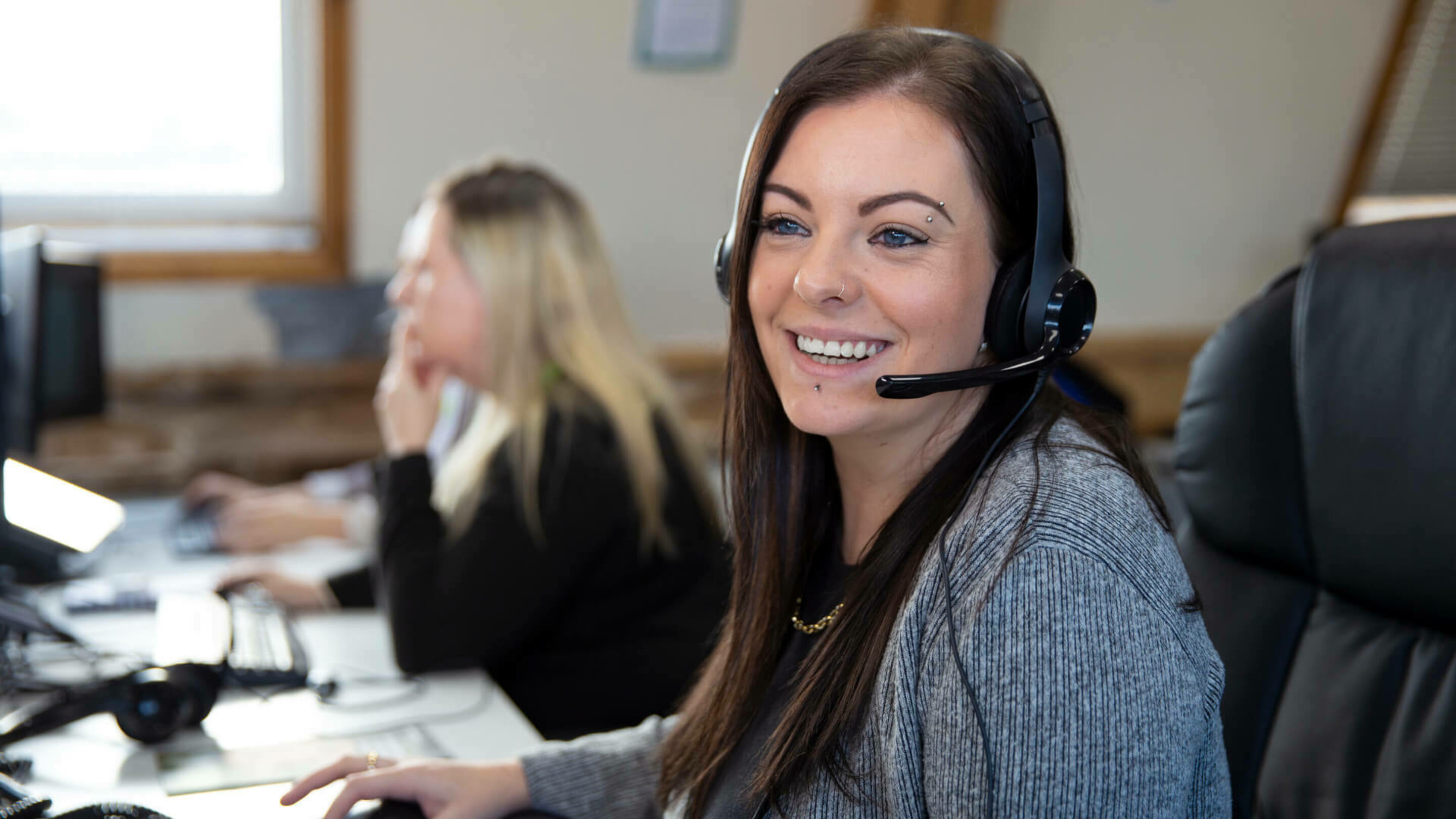 Two women looking at computer screens, wearing phone headsets and smiling.