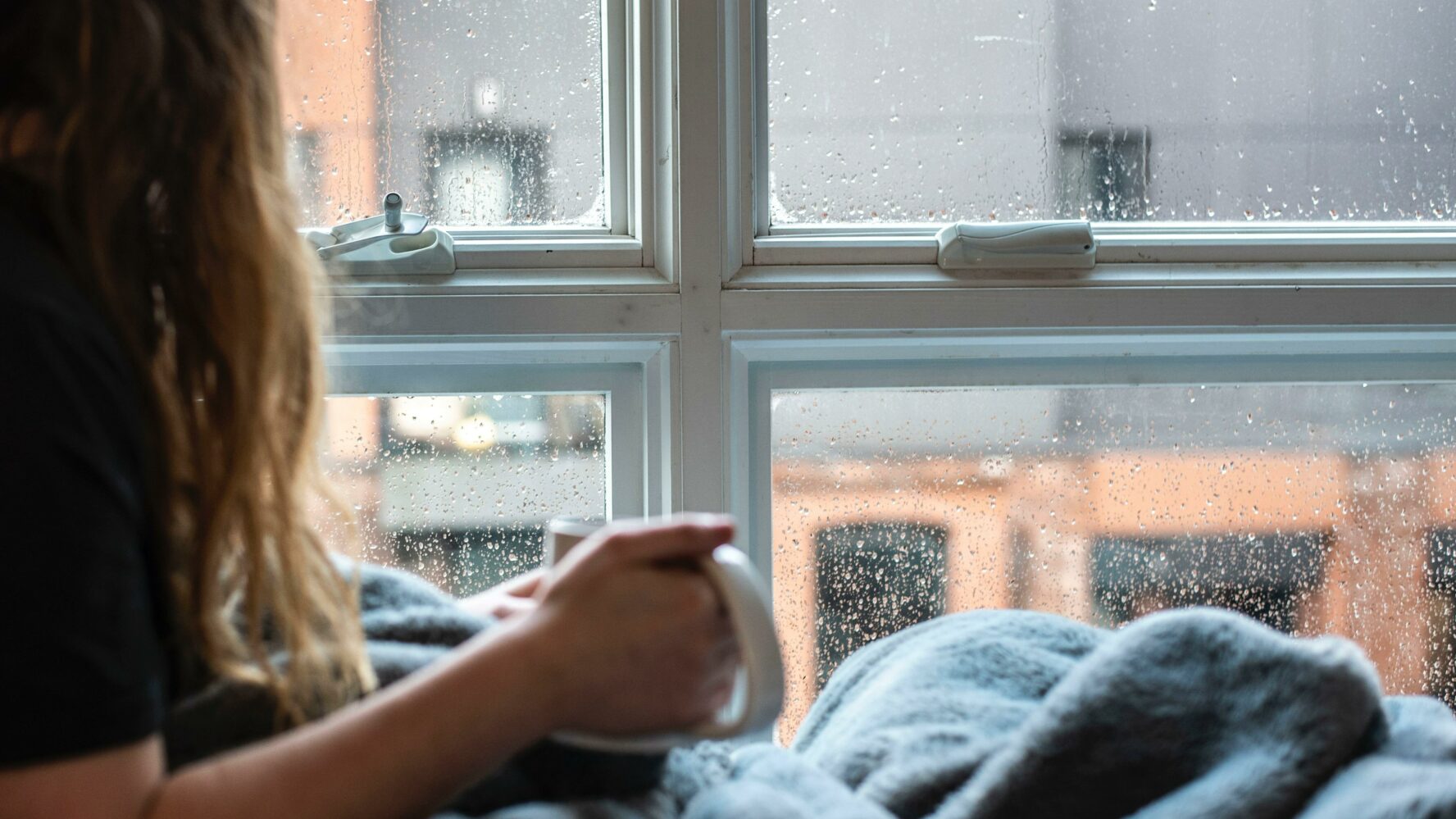 Woman sat at window, in a blanket with hot drink