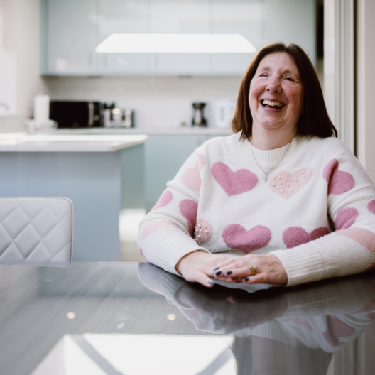 Angela, a white female with brown, shoulder length hair, wearing a fluffy white jumper with pink hearts. She is sat at her kitchen table, smiling at the camera.