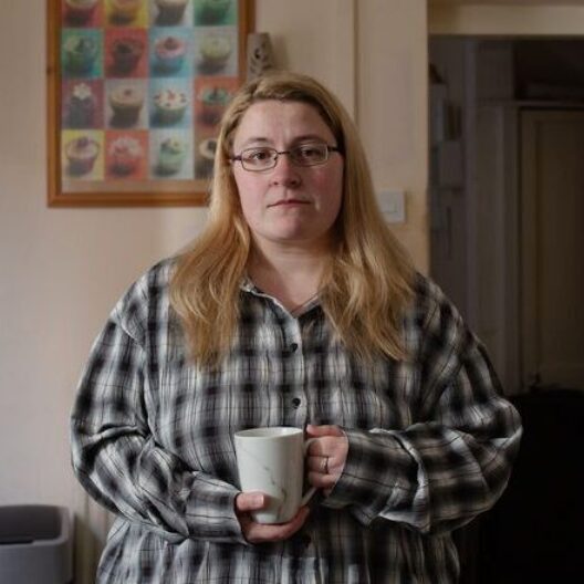 Nicole, a former CAP client, wearing a black and white checked shirt, looking at the camera with a serious face and a mug of tea in her hand.