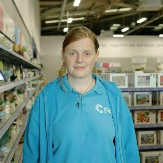 CAP client, Lyndsey, stood in a charity shop where she volunteers, smiling at the camera