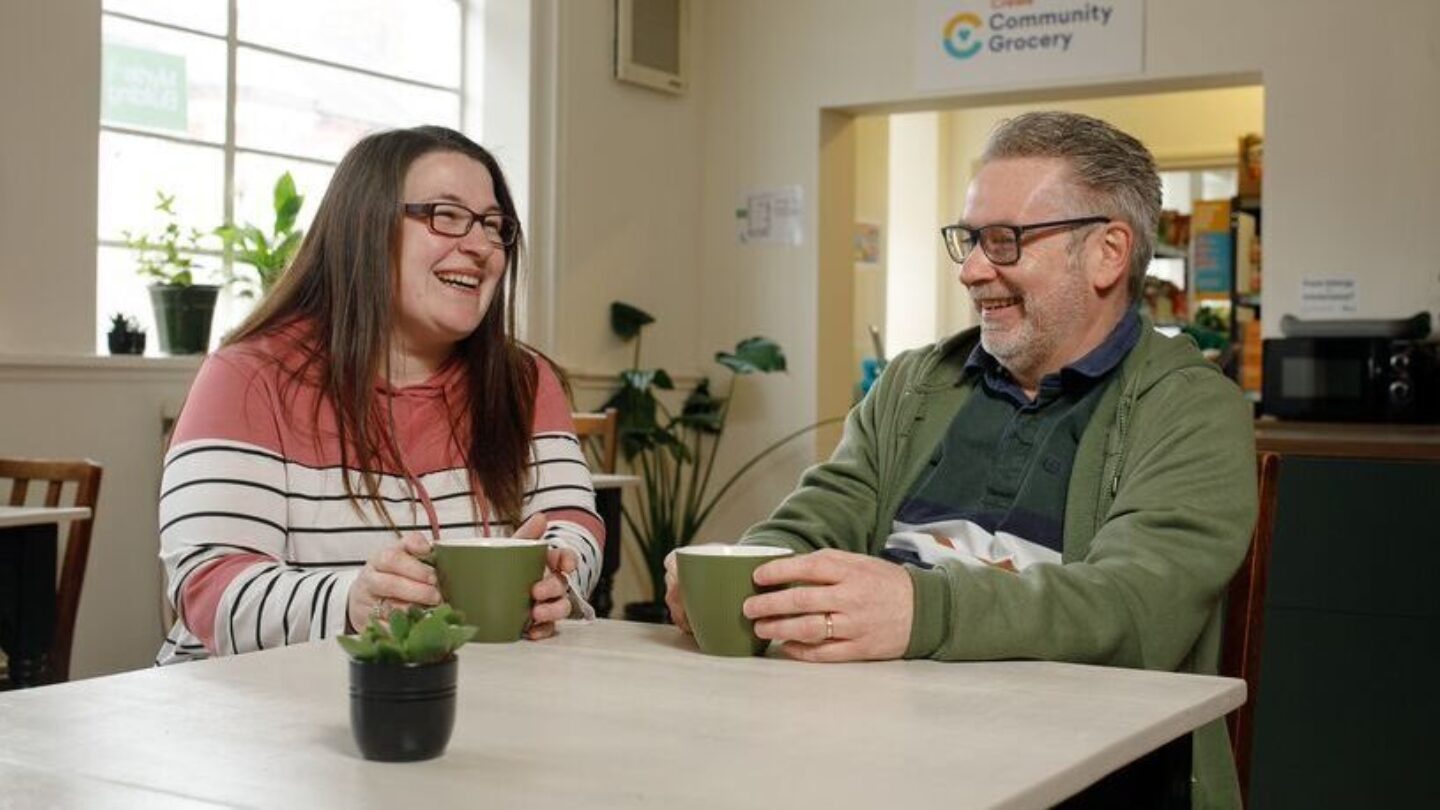 Former CAP client Nicole, a white lady with brown straight hair and glasses, sat next to Debt Coach Tim, a white male with grey hair and glasses. They're both having a cup of tea together at the table of their local church-based community cafe.