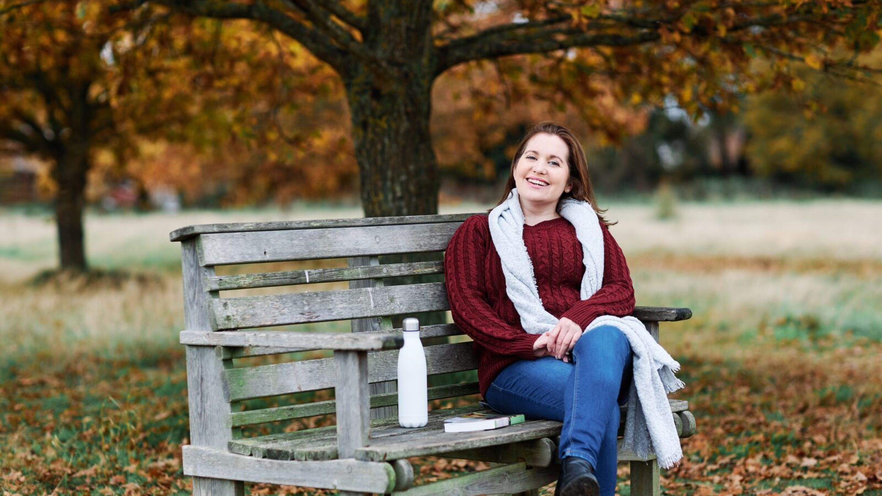 CAP client, Charlotte, smiling and sat on a park bench in autumn, wearing a jumper and a scarf