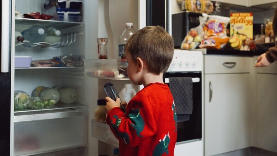 Young child wearing a red Christmas jumper and looking in his fridge, holding a grocery item