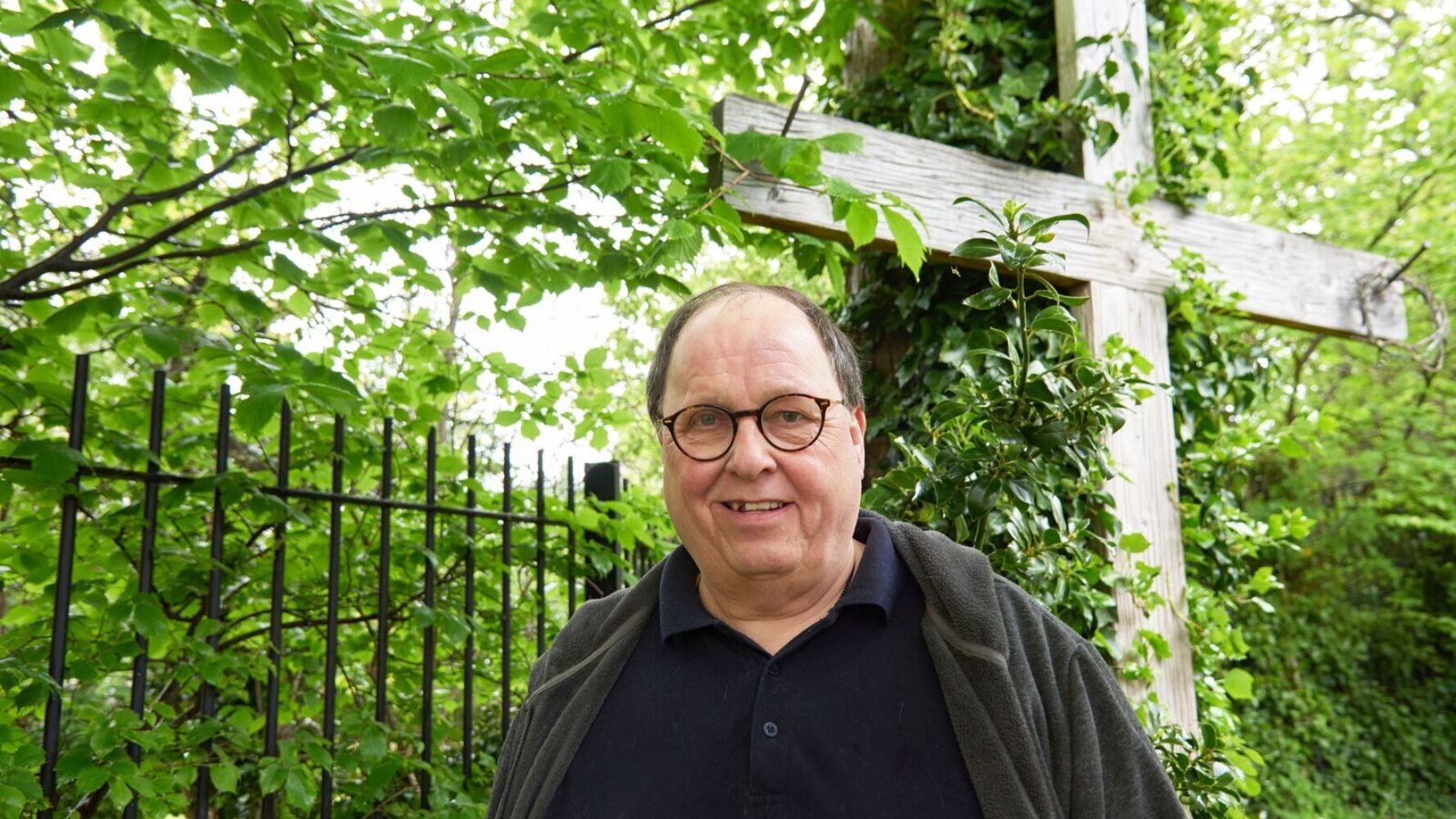 CAP client, Simon, stood outside in a garden, smiling, with a large wooden cross propped up against a tree behind him