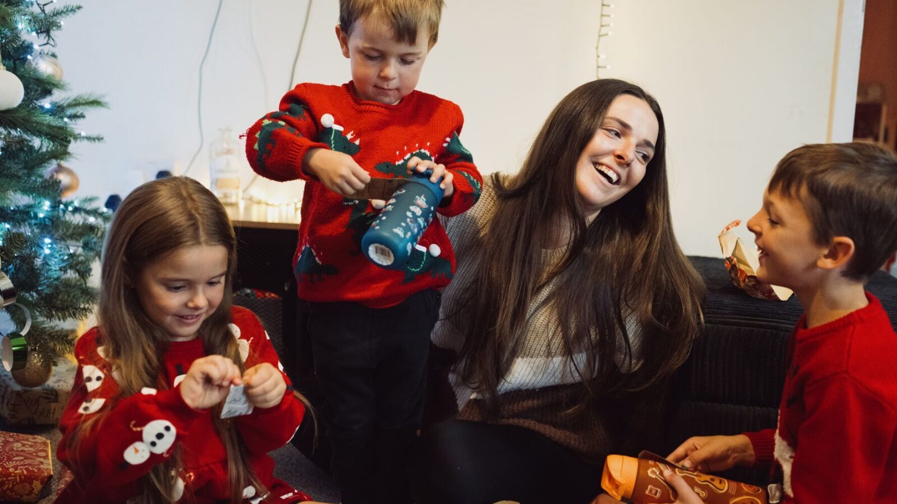 CAP client, Jade, sat with her three children in her living room next to a Christmas tree. The three children are all wearing red Christmas jumpers and are opening presents.