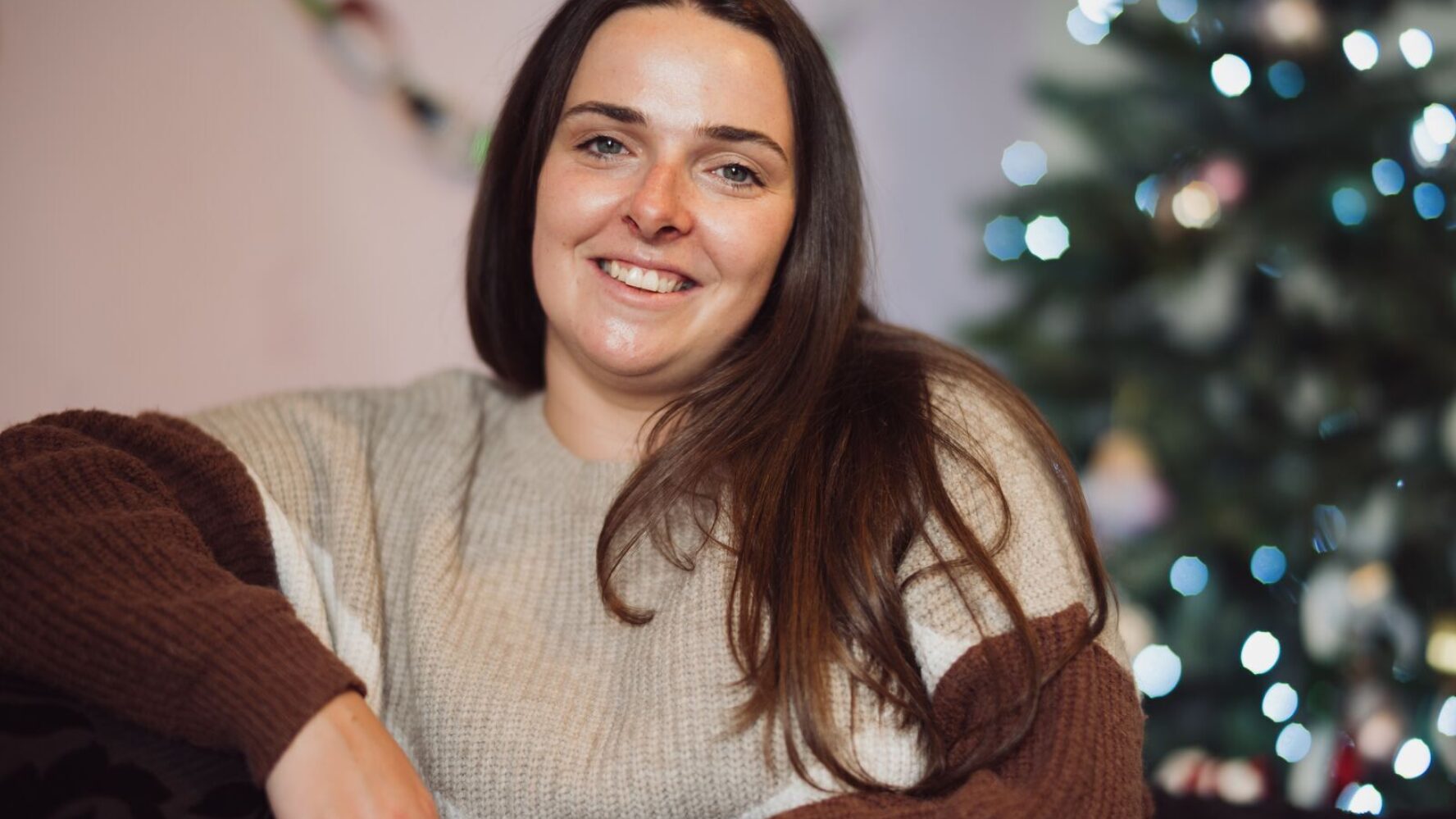 CAP client, Jade, sat in her living room smiling at the camera, with her Christmas tree and decorations behind her