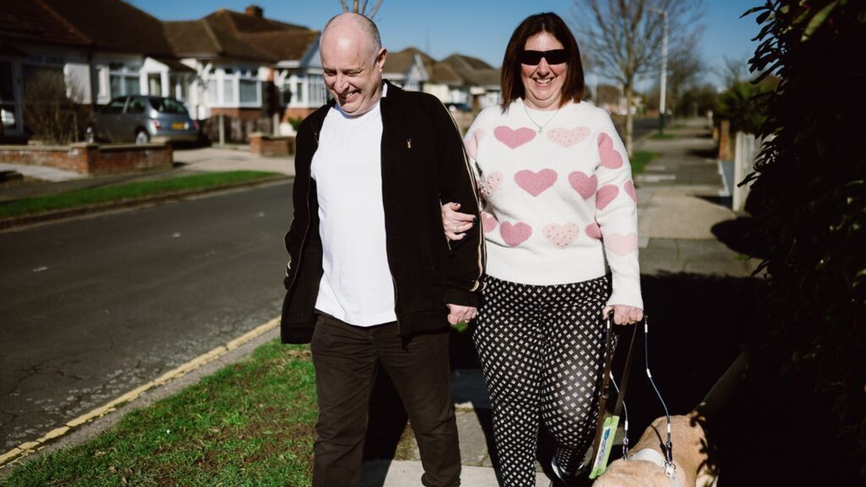 Picture of a husband and wife walking arm in arm down their street with their guide dog